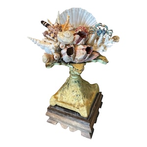 May include: A decorative pedestal stand featuring a square wooden base and a distressed yellow column topped with an arrangement of various seashells, white coral fragments, and a small blue rhinestone-encrusted crab brooch ornament.