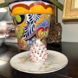 May include: A white porcelain pomegranate figurine adorned with small pink sculpted roses and gold-painted accents, resting on a matching white porcelain saucer featuring a delicate floral pattern with a thin gold rim along the edge.