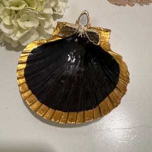 May include: A decorative seashell-shaped trinket dish. The dish is black with a gold-painted rim and a gold bow-shaped embellishment with a central gem. The dish is approximately 6 inches in diameter, perfect for holding small jewelry items.