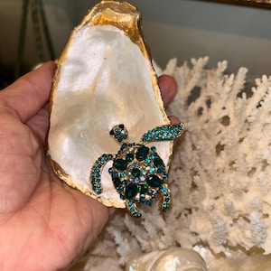 May include: A decorative oyster shell with a gold-painted edge, holding a bejeweled turtle figurine. The turtle features green and blue crystals. The shell is white and iridescent, and the background includes coral and a seashell.