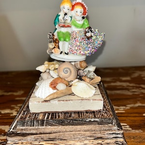 Peut inclure: Une figurine décorative représentant deux personnages en porcelaine vêtus de vert et de blanc, la figure féminine portant une jupe recouverte de strass irisés. La base est ornée de divers coquillages naturels sur un support en bois à étages.