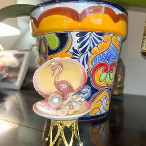 May include: A decorative seashell ornament featuring a glittery pink flamingo figurine with a small crown, a large white faux pearl, and small clear crystals. The shell is mounted on a gold-toned metal stand with an intricate arched base design.