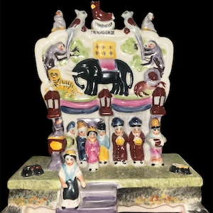 May include: A detailed ceramic figurine depicting a menagerie scene. The figurine features an elephant, monkeys, birds, and other animals. The word "MENAGERIE" is visible. The base includes figures of people and a woman carrying a bag.
