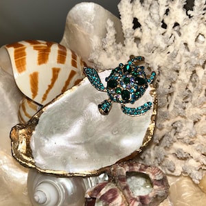 May include: A jeweled turtle brooch with emerald and teal stones rests on a pearl oyster shell with a gold rim. The shell is surrounded by other seashells and coral, creating a coastal theme.