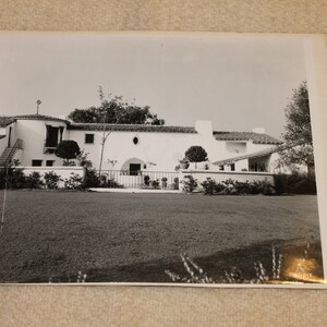 May include: A black and white photograph of a white, Spanish-style house with a tiled roof. The house has a large front yard with a lawn and bushes.