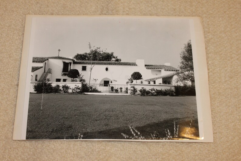 May include: A black and white photograph of a white, Spanish-style house with a tiled roof. The house has a large front yard with a lawn and bushes.