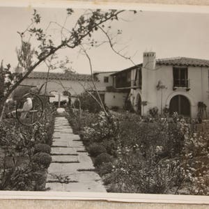May include: A black and white photograph of a Spanish-style house with a garden in the foreground. The house has a white stucco exterior and a red tile roof. The garden is filled with lush greenery and flowers.