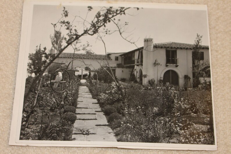 May include: A black and white photograph of a Spanish-style house with a garden in the foreground. The house has a white stucco exterior and a red tile roof. The garden is filled with lush greenery and flowers.