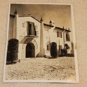 May include: A black and white photograph of a large, white, Spanish-style house with a tiled roof. The house has a large, arched doorway and a balcony with a wrought iron railing. There are trees and bushes in the front yard.