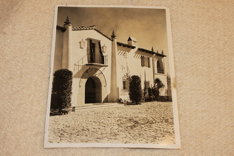 May include: A black and white photograph of a large, white, Spanish-style house with a tiled roof. The house has a large, arched doorway and a balcony with a wrought iron railing. There are trees and bushes in the front yard.