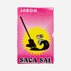 Puede incluir: Una caja rosa con las palabras "Jabon Saca Sal" impresas en ella. La caja presenta una ilustración en negro y amarillo de una escoba que barre un montón de sal.