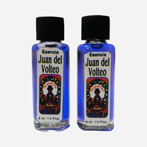 May include: Two small blue glass bottles with black caps. Each bottle contains a clear liquid and has a white label with the text "Esencia Juan del Volteo" and a colorful illustration of a man in a hat. The label also includes the text "8 ml. 1/4 Floz."