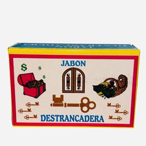 May include: A yellow, red, and blue box with the words "Jabon Destrancadera" printed on it. The box features illustrations of a key, a chest full of money, a door, and a cornucopia.