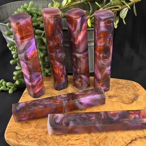May include: A collection of six rectangular resin blocks with a marbled effect. The blocks feature a mix of purple, red, and orange hues. The blocks are displayed on a wooden cutting board, with a metal bucket and greenery in the background.