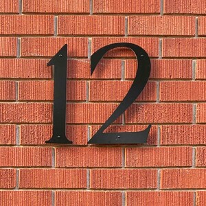 May include: Black metal house number "12" mounted on a red brick wall. The numbers are in a stylized font and are attached to the wall with small screws. The brick has a textured surface.