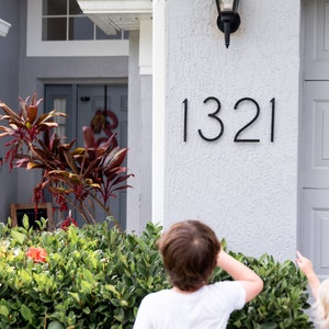 THIN MODERN Black house numbers 1321 mounted horizontally on grey stucco beside the garage door. 2 children are pointing toward the new numbers.