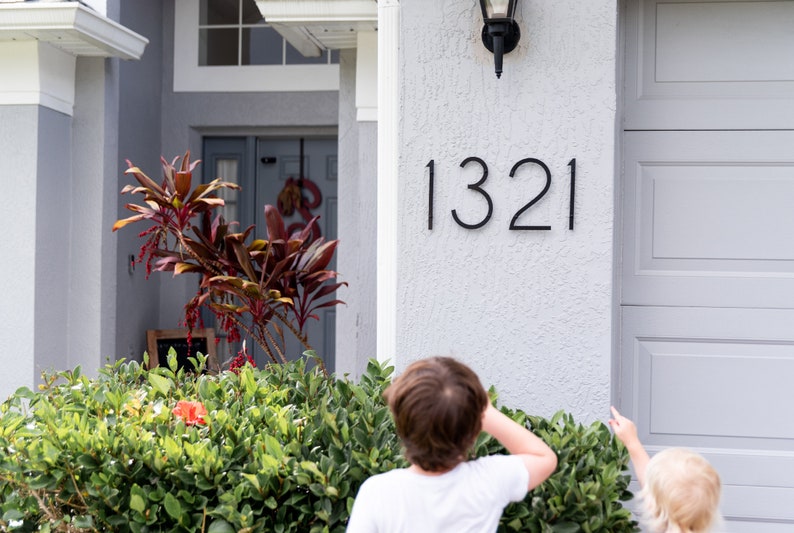 THIN MODERN Black house numbers 1321 mounted horizontally on grey stucco beside the garage door. 2 children are pointing toward the new numbers.