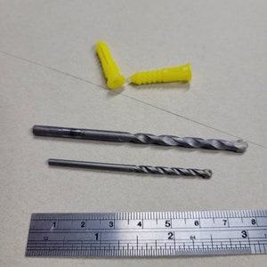 May include: Two yellow plastic wall anchors and two drill bits, one longer than the other, are laid out on a white surface next to a metal ruler marked in inches.