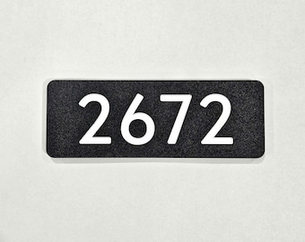 Puede incluir: Placa de dirección rectangular negra con el número "2672" en blanco. Los números están centrados y en una fuente clara sin serifa. La placa tiene esquinas redondeadas y una superficie texturizada.
