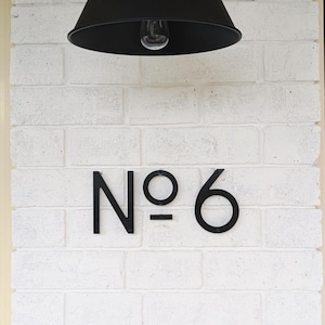A modern house number sign with 5 inch THIN MODERN No letters and numbers fastened with screws and spacers onto white painted brick.