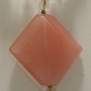 May include: A pink gemstone pendant necklace with a large, faceted, pink gemstone pendant. The pendant is attached to a chain of smaller, clear and pink gemstones. The necklace has a gold-toned clasp and a gold-toned spiral charm.