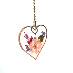 May include: A heart-shaped pendant with a gold chain, featuring pressed flowers in shades of pink, purple, and green encased in clear resin.
