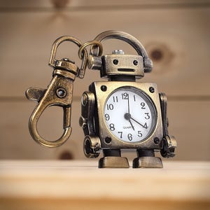 May include: A bronze-toned robot-shaped keychain watch. The watch face is white with black numerals and hands. The robot has a square body, round head, and a clasp for attaching to keys or a bag. The background is a wooden surface.