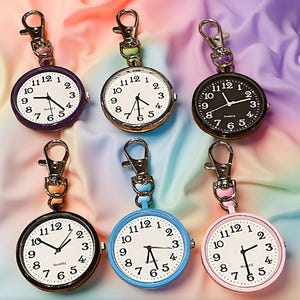 Op de afbeelding: Zes zakhorloges met quartz uurwerken en metalen sluitingen. De horloges hebben witte wijzerplaten met zwarte cijfers en wijzers. De horlogekasten zijn in verschillende kleuren: paars, goud, zwart, zwart, blauw en roze. De achtergrond is een kleurrijke, gedrapeerde stof.