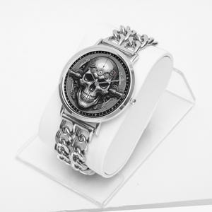 A silver wristwatch with a black face featuring a detailed skull design. The watch has a silver chain link band.