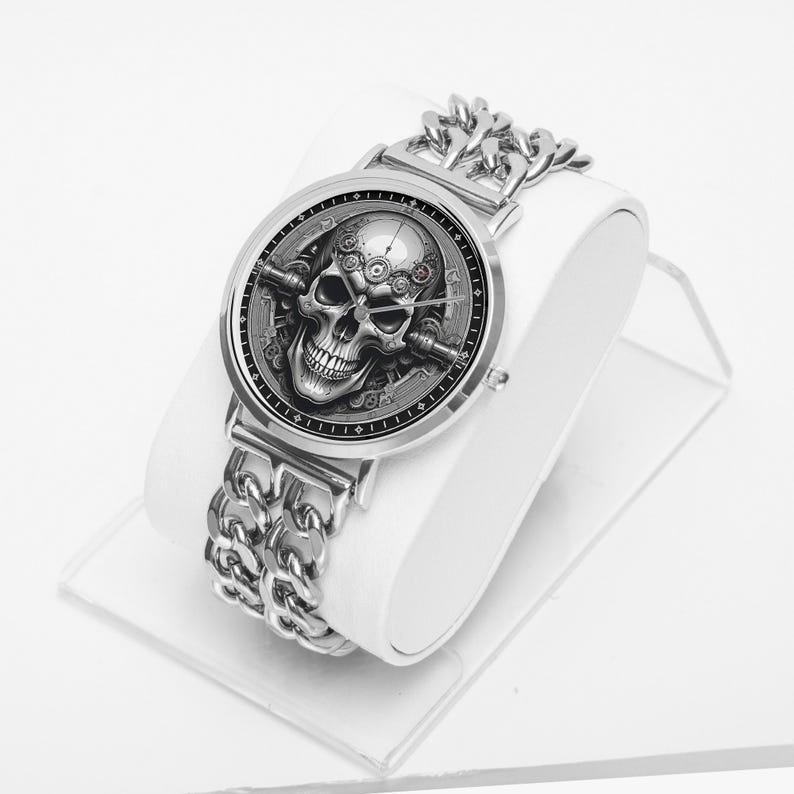 A silver wristwatch with a black face featuring a detailed skull design. The watch has a silver chain link band.