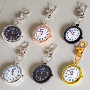 Puede incluir: Una colección de relojes de bolsillo de cuarzo con varios acabados metálicos, incluyendo plata, oro y negro. Cada reloj tiene una esfera blanca o negra con números arábigos y un clip.
