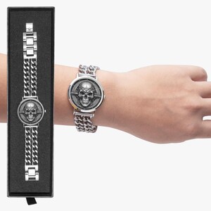A silver watch with a skull design on the face and a chain link band. The watch is in a black box.