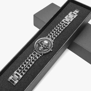 A silver watch with a skull and crossbones design on the face. The watch is in a black box with a black velvet lining.