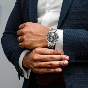 A silver watch with a skull design on the face and a silver chain link band.