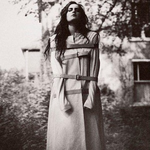 May include: Monochrome image of a person wearing a long, light-colored coat with multiple leather straps. The person is standing outdoors with a blurred background of foliage and a building. The coat appears to be a straightjacket.
