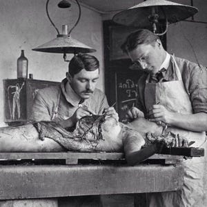 May include: Black and white vintage photograph of two men performing a medical procedure on a body. They are wearing aprons and working under a lamp. A chalkboard with writing is in the background.