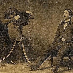 May include: A black and white photograph of a man sitting in a chair while a person takes his picture with a large camera on a tripod.