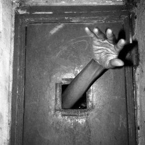 May include: A black and white photograph of a metal door with the text NO. 5 printed above a small square opening. An arm and hand extend outward through the opening, with the fingers spread wide against the dark, textured surface of the door.