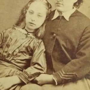 May include: A black and white vintage photograph of two women. The woman on the left is wearing a long-sleeved dress and has her head resting on the shoulder of the woman on the right. The woman on the right is wearing a dark jacket and has her hair pulled back.