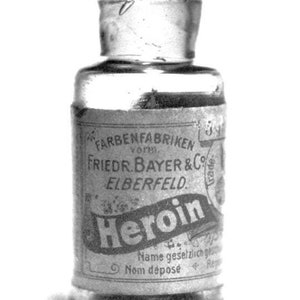 May include: A small, clear glass bottle with a metal cap. The label reads "Heroin" in a stylized font, with additional text indicating the manufacturer, "Friedr. Bayer & Co Elberfeld."