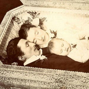 May include: Vintage sepia photograph showing three people in a coffin. Two adults and a baby are lying together, surrounded by flowers. The coffin has decorative trim.
