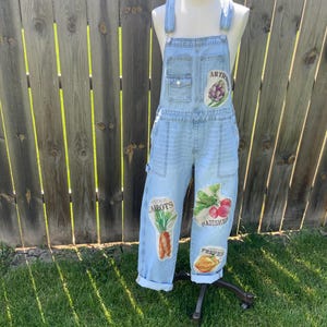 May include: Light blue denim overalls featuring a chest pocket and multiple vegetable-themed patches. The patches display illustrations of an artichoke, carrots, radishes, and peppers with the text Artichokes, Heritage Carrots, Radishes, and Peppers.