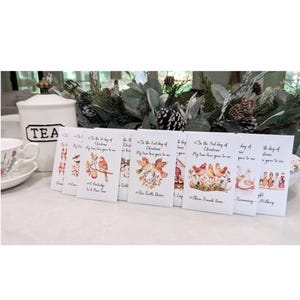 May include: A collection of Christmas cards with watercolor illustrations. Each card features a different design, including birds, swans, and people, with text from the "12 Days of Christmas" song. The cards are arranged on a white surface.