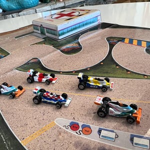 May include: A toy race track mat featuring a printed hospital building with a red cross symbol and five miniature formula-style race cars in various colors including white, red, blue, yellow, and orange, positioned on the track surface.
