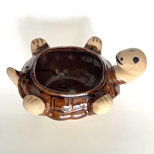 May include: A ceramic turtle-shaped planter with a glossy brown finish. The turtle has a light tan head, feet, and tail, with black dot eyes and a smiling mouth. The planter's opening is a large, open space.