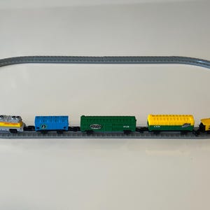 May include: A toy train set consisting of a gray oval track and five connected train cars. The train includes a yellow and gray locomotive, a blue boxcar, a green freight car, a yellow and green hopper car, and a small yellow caboose.