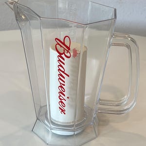 May include: A clear, faceted plastic pitcher featuring a central white cylindrical insert with the red Budweiser logo and a small red crown icon printed on the front. The pitcher has a sturdy handle and a molded spout for pouring liquids.