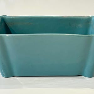 May include: A rectangular turquoise ceramic planter with a smooth, glossy finish. The container features slightly flared sides and rounded corners, with a wide, open top and a solid base, presenting a clean and simple design for holding items.