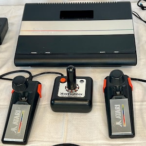 May include: A black Atari video game console with two joysticks and a StarFighter joystick. The console has a silver stripe with red, green, and blue accents. The joysticks are black with orange accents and the Atari logo.