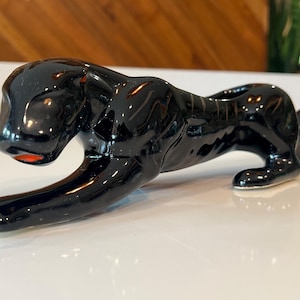 May include: A glossy black ceramic panther figurine in a low, prowling stance. The sculpture features a smooth, reflective surface with a distinct orange accent on the mouth area, capturing the animal in a stretched, elongated pose.